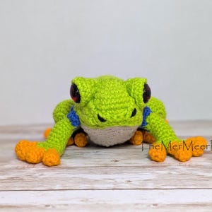 May include: A crocheted frog toy featuring a bright lime green body, a white underbelly, blue accents on the upper limbs, and vibrant orange feet. The frog has large, dark, glossy eyes and the text TheMerMeerKat is printed on the right side.