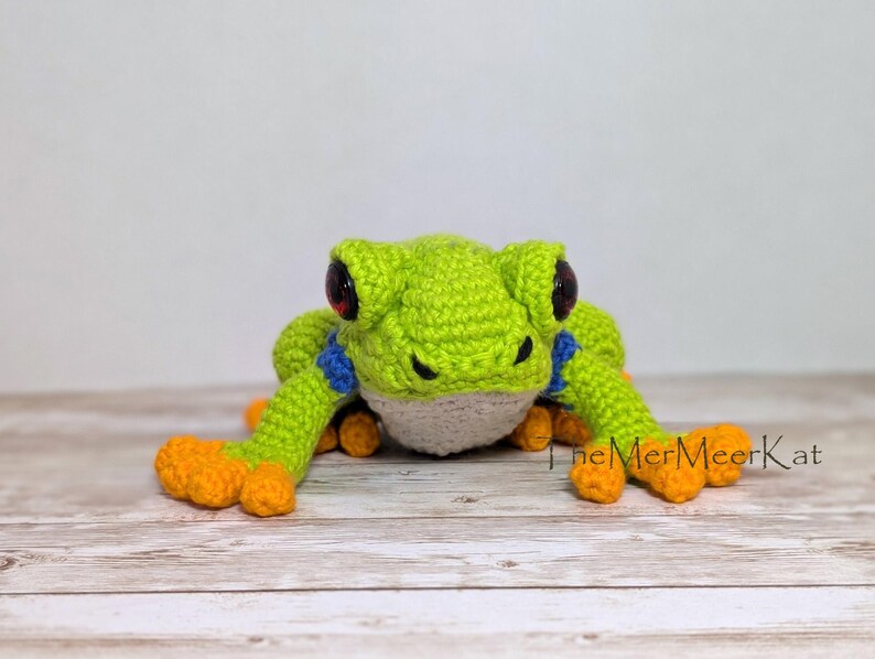 May include: A crocheted frog toy featuring a bright lime green body, a white underbelly, blue accents on the upper limbs, and vibrant orange feet. The frog has large, dark, glossy eyes and the text TheMerMeerKat is printed on the right side.