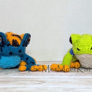 May include: Two crocheted frog figures. The left frog is blue with black spots and orange accents on its head and feet. The right frog is lime green with a white belly, blue bands on its front legs, and orange feet. Both have large dark eyes.