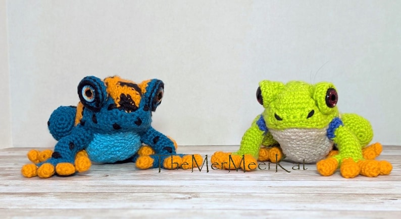 May include: Two crocheted frog figures. The left frog is blue with black spots and orange accents on its head and feet. The right frog is lime green with a white belly, blue bands on its front legs, and orange feet. Both have large dark eyes.