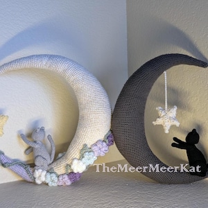 Pode incluir: Duas decorações de meia-lua de crochê, uma branca e outra cinza. Cada lua tem uma estrela pendurada e um gato. A lua branca tem um gato cinza e flores em tons pastel. A lua cinza tem um gato preto. Texto TheMeerMeerKat visível.