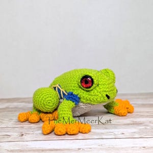 May include: A crocheted frog toy with a bright green body, orange feet, and large red eyes. The frog features blue and yellow accents on its sides and a white underbelly. The text TheMerMeerKat is printed in black near the base of the frog.