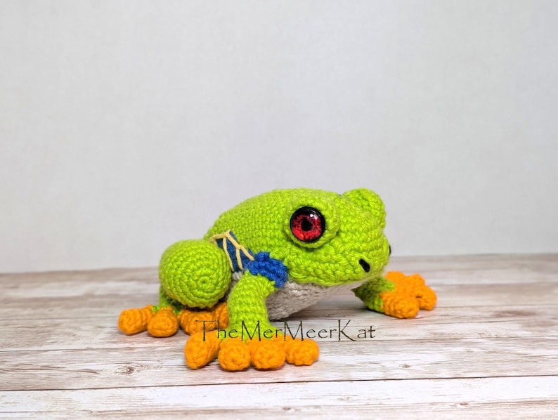 May include: A crocheted frog toy with a bright green body, orange feet, and large red eyes. The frog features blue and yellow accents on its sides and a white underbelly. The text TheMerMeerKat is printed in black near the base of the frog.
