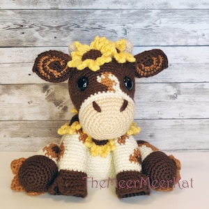 May include: A crocheted brown and white cow with a yellow sunflower on its head. The cow is sitting on a white surface.