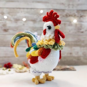 May include: A handcrafted crochet rooster figure. The rooster features a white body, a red comb and wattle, and a colourful tail. The neck is decorated with a yellow, green, and red collar. The rooster's feet are yellow. The MeerMeerKat is written on the bottom.