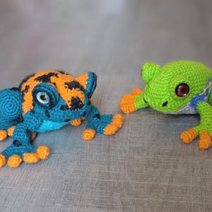May include: Two handmade crochet frogs. The left frog features a teal body with black spots, orange patches on its back, and orange toes. The right frog is bright green with a blue and white side pattern, dark red eyes, and orange toes.