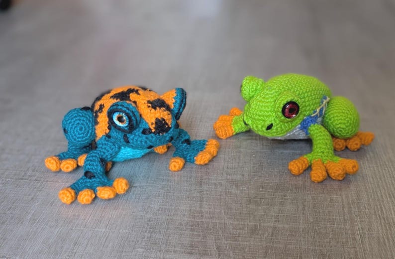 May include: Two handmade crochet frogs. The left frog features a teal body with black spots, orange patches on its back, and orange toes. The right frog is bright green with a blue and white side pattern, dark red eyes, and orange toes.