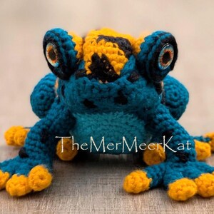 May include: A crocheted frog toy featuring a teal body with black spots, a yellow head patch, and bright yellow toes. The frog has large, round, multi-colored eyes and the text TheMerMeerKat is printed in white across the center of the body.