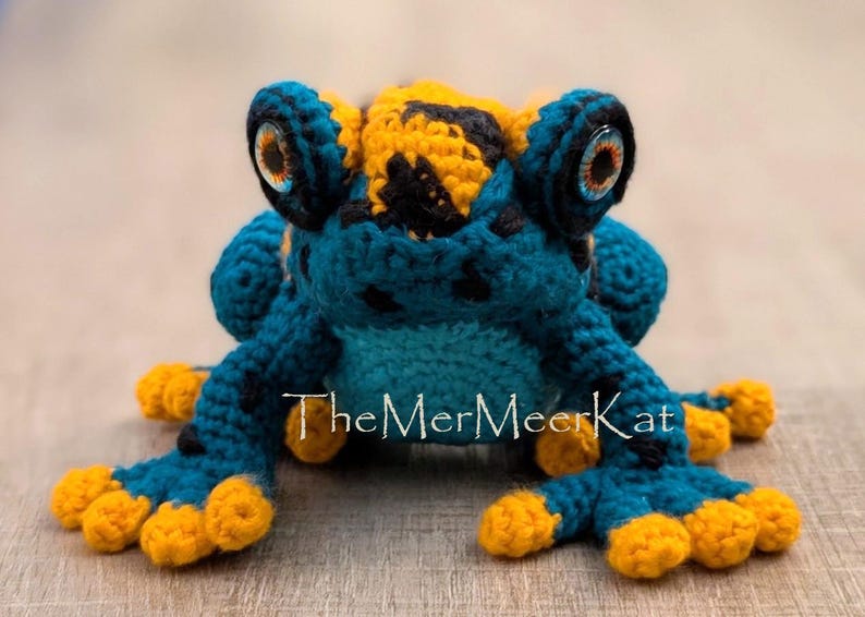 May include: A crocheted frog toy featuring a teal body with black spots, a yellow head patch, and bright yellow toes. The frog has large, round, multi-colored eyes and the text TheMerMeerKat is printed in white across the center of the body.
