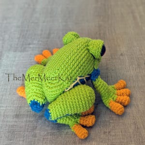 May include: A crocheted frog toy featuring a bright green body, orange toes, and blue accents on the legs and sides. The text TheMerMeerKat is printed in black on the left side of the frog. The frog has dark, round eyes and a textured surface.