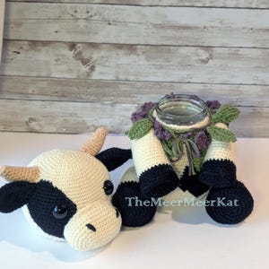 Puede incluir: Funda de tarro y cabeza de vaca de ganchillo con temática de vaca. La funda del tarro es blanca con detalles en negro, flores moradas y hojas verdes. La cabeza de la vaca es blanca con manchas negras y cuernos color canela. El tarro tiene el texto "TheMeerMeerKat".