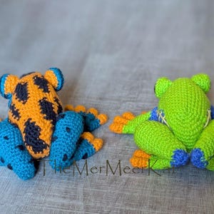 May include: Two crocheted frog toys. The left frog is orange with black spots and blue limbs with black spots. The right frog is bright green with blue accents on the limbs and orange toes. The text TheMerMeerKat is visible at the bottom center.
