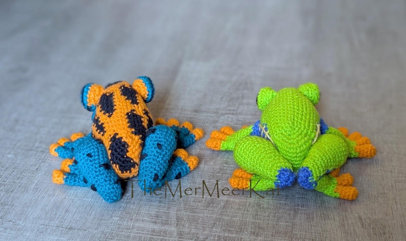 May include: Two crocheted frog toys. The left frog is orange with black spots and blue limbs with black spots. The right frog is bright green with blue accents on the limbs and orange toes. The text TheMerMeerKat is visible at the bottom center.