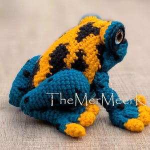 May include: A crocheted frog toy featuring a bright yellow back with black spots and deep blue limbs with yellow-tipped toes. The frog has a realistic-looking eye with a brown iris and black pupil. The text TheMerMeerKat is visible.
