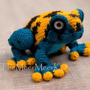 May include: A crocheted frog toy featuring a vibrant yellow back with black spots, a deep blue head and limbs, and bright yellow toes. The frog has large, detailed eyes with orange irises. The text TheMeerMeerKat is printed in white.