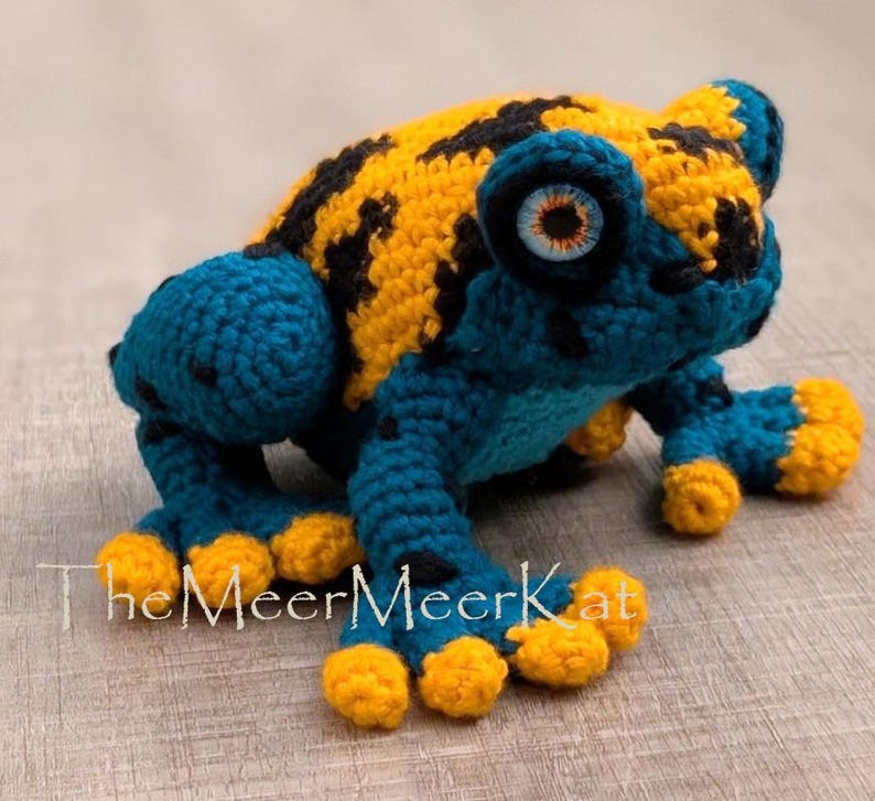 May include: A crocheted frog toy featuring a vibrant yellow back with black spots, a deep blue head and limbs, and bright yellow toes. The frog has large, detailed eyes with orange irises. The text TheMeerMeerKat is printed in white.