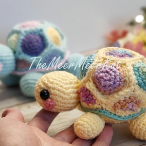 May include: Two crocheted turtle toys. One is light blue with pastel pink, yellow, and purple accents. The other is yellow with pastel pink, purple, and blue accents. The Meer Meer Knot logo is visible on the blue turtle.