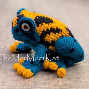 May include: A crocheted frog figurine featuring a vibrant yellow and black spotted back, with deep blue limbs and head. The frog has large, detailed eyes with orange irises and blue pupils. The text TheMerMeerKat is visible on the lower left side.