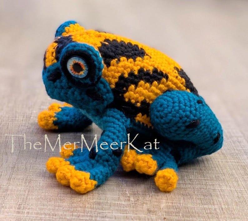 May include: A crocheted frog figurine featuring a vibrant yellow and black spotted back, with deep blue limbs and head. The frog has large, detailed eyes with orange irises and blue pupils. The text TheMerMeerKat is visible on the lower left side.