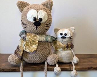 May include: Two crocheted cat plushies with large white eyes, black pupils, and small black noses. The larger cat is light brown, and the smaller cat is beige. Both cats wear matching variegated yellow, brown, and green crocheted scarves.