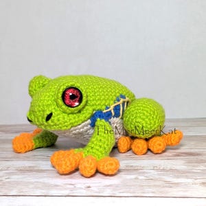 May include: A crocheted frog toy featuring a bright green body, orange feet, and large red eyes. The frog has blue and yellow stitched details on its side and a cream-colored belly. The text TheMerMeerKat is visible near the frog.