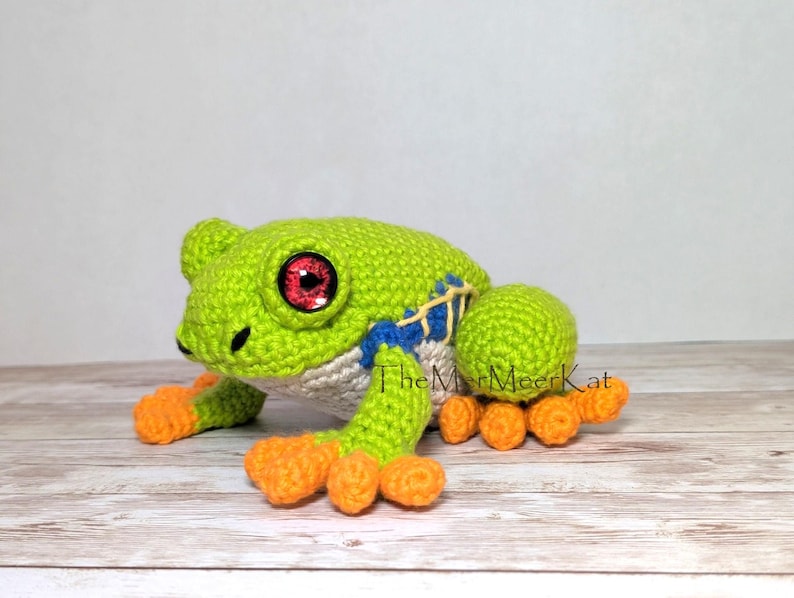 May include: A crocheted frog toy featuring a bright green body, orange feet, and large red eyes. The frog has blue and yellow stitched details on its side and a cream-colored belly. The text TheMerMeerKat is visible near the frog.
