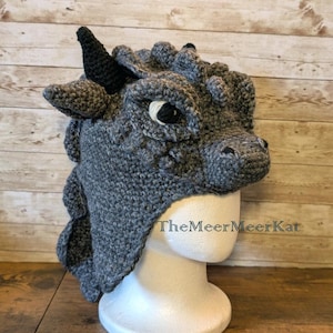 May include: A grey crocheted dragon hat with black horns and eyes, displayed on a white mannequin head. The hat features the text "TheMeerMeerKat". The hat is made from soft, textured yarn. The hat is a unique, handmade item.