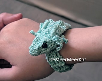 May include: A light mint green crochet bracelet featuring a small dragon head with black bead eyes and textured horns. The dragon is integrated into the band, which has an open-work pattern. The text TheMeerMeerKat is printed on the image.