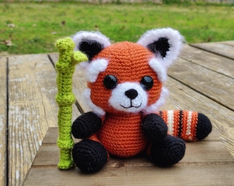 May include: A crocheted red panda stuffed animal with orange, white, and black yarn, featuring large black safety eyes and a small black nose. Beside the red panda is a tall, segmented green crocheted bamboo stalk.