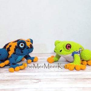 May include: Two crocheted frogs. The left frog is blue and orange with black spots and blue eyes. The right frog is light green with a white belly, blue side markings, and red eyes. The text TheMerMeerKat is printed between the two frogs.
