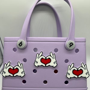 Peut inclure: Un sac fourre-tout de couleur lavande avec un logo "bogg". Le sac a deux boutons blancs avec des num&eacute;ros noirs. Il y a trois breloques en forme de main blanche avec des c&oelig;urs rouges.