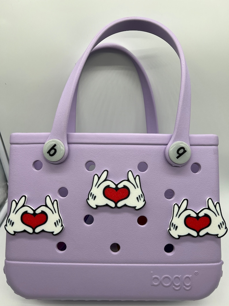 Peut inclure: Un sac fourre-tout de couleur lavande avec un logo "bogg". Le sac a deux boutons blancs avec des num&eacute;ros noirs. Il y a trois breloques en forme de main blanche avec des c&oelig;urs rouges.
