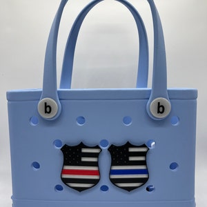 Puede incluir: Una bolsa de mano de color azul claro con dos parches en forma de escudo de color negro y blanco. Los parches muestran la bandera estadounidense con una franja roja y una franja azul, que representan la Línea Roja Fina y la Línea Azul Fina.