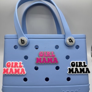 Puede incluir: Una bolsa de mano azul claro con la palabra "bogg" impresa en la parte inferior. La bolsa tiene tres abalorios "Girl Mama" unidos a ella. Un abalorio es rosa y blanco, otro es negro y blanco, y otro es rosa, blanco y gris.