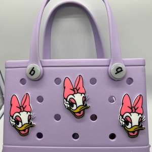 Peut inclure: Un sac fourre-tout de couleur lavande avec un logo "bogg". Le sac a trois personnages de canard de dessin anim&eacute; avec des n&oelig;uds roses attach&eacute;s.