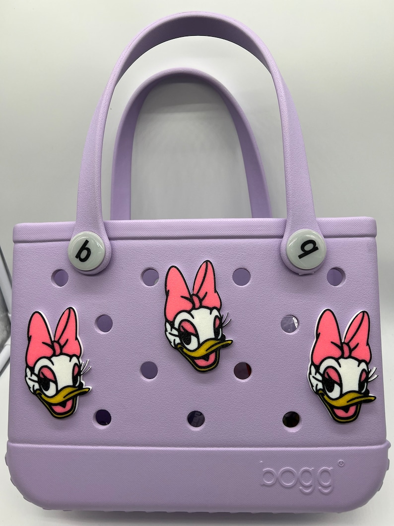 Peut inclure: Un sac fourre-tout de couleur lavande avec un logo "bogg". Le sac a trois personnages de canard de dessin anim&eacute; avec des n&oelig;uds roses attach&eacute;s.
