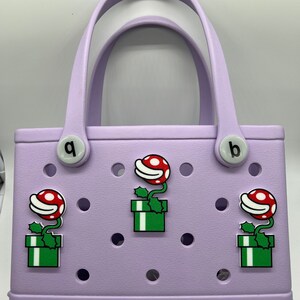 Puede incluir: Un bolso tote lila con dos asas y la palabra "bogg" impresa en la parte inferior. El bolso est&aacute; decorado con tres dijes de plantas de dibujos animados rojos y blancos en cajas verdes. El bolso tiene dos dijes circulares blancos con las letras "q" y "b".