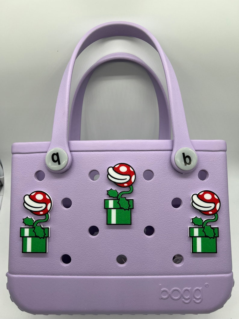 Puede incluir: Un bolso tote lila con dos asas y la palabra "bogg" impresa en la parte inferior. El bolso est&aacute; decorado con tres dijes de plantas de dibujos animados rojos y blancos en cajas verdes. El bolso tiene dos dijes circulares blancos con las letras "q" y "b".