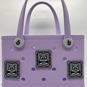 Puede incluir: Un bolso tote lavanda con dos asas y la letra "b" a cada lado. El bolso presenta tres parches decorativos cuadrados grises con un dise&ntilde;o de cara de dibujos animados. La palabra "bogg" est&aacute; impresa en la parte inferior.