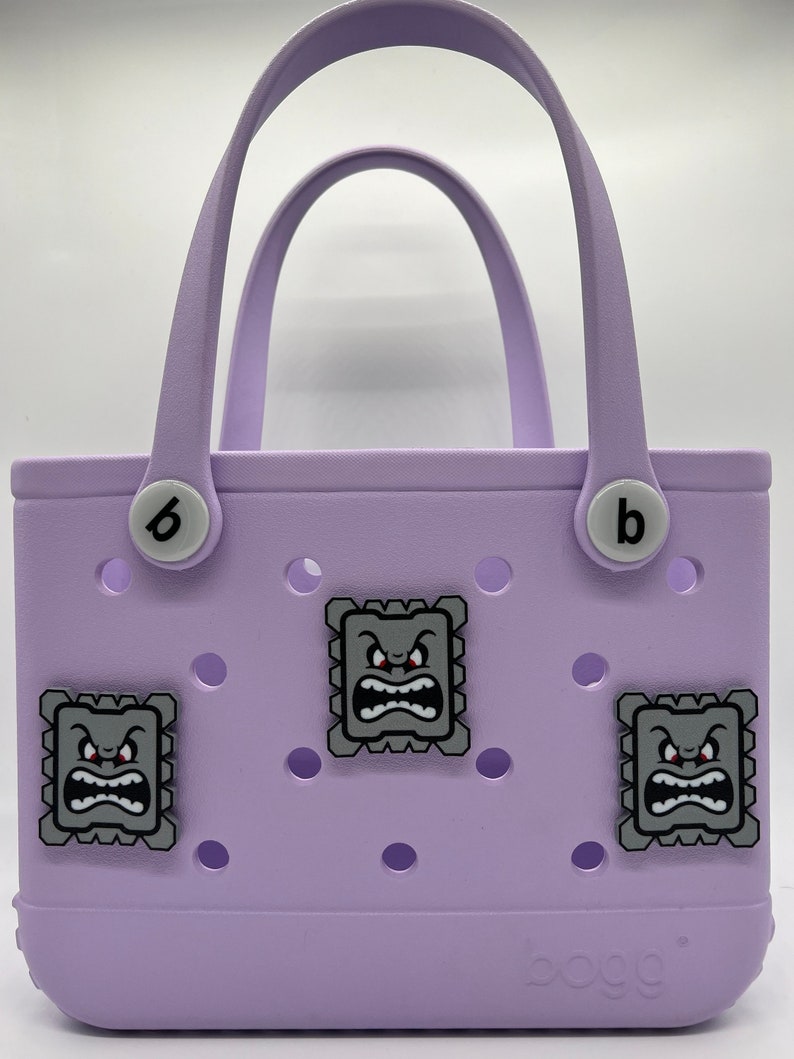 Puede incluir: Un bolso tote lavanda con dos asas y la letra "b" a cada lado. El bolso presenta tres parches decorativos cuadrados grises con un dise&ntilde;o de cara de dibujos animados. La palabra "bogg" est&aacute; impresa en la parte inferior.