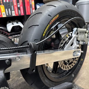 Könnte beinhalten: Nahaufnahme eines schwarzen Motorradreifens mit dem Text "KENDA CURVE" in Gelb. Der Reifen ist auf einer silbernen Felge mit einer Scheibenbremse montiert. Das Bild zeigt einen Teil des Rahmens und der Federung des Motorrads.