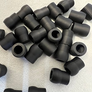 May include: A collection of small, black rubber or plastic cylindrical components with a flared shape. The components have a hollow center and are scattered across a light-colored surface. The objects appear to be new and unused.