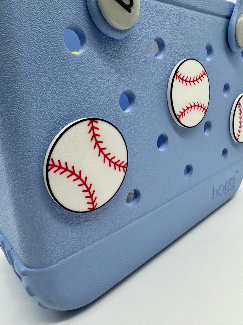 Peut inclure: Un sac fourre-tout bleu clair orn&eacute; de breloques sur le th&egrave;me du baseball. Les breloques sont blanches avec des coutures rouges, ressemblant &agrave; des balles de baseball. Le sac a une surface textur&eacute;e et plusieurs trous ronds.