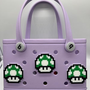 Puede incluir: Un bolso tote lavanda con asas blancas y adornos en forma de hongo. La bolsa tiene la palabra "bogg" impresa en la esquina inferior derecha. Tiene dos botones blancos con las letras "b" y "g".