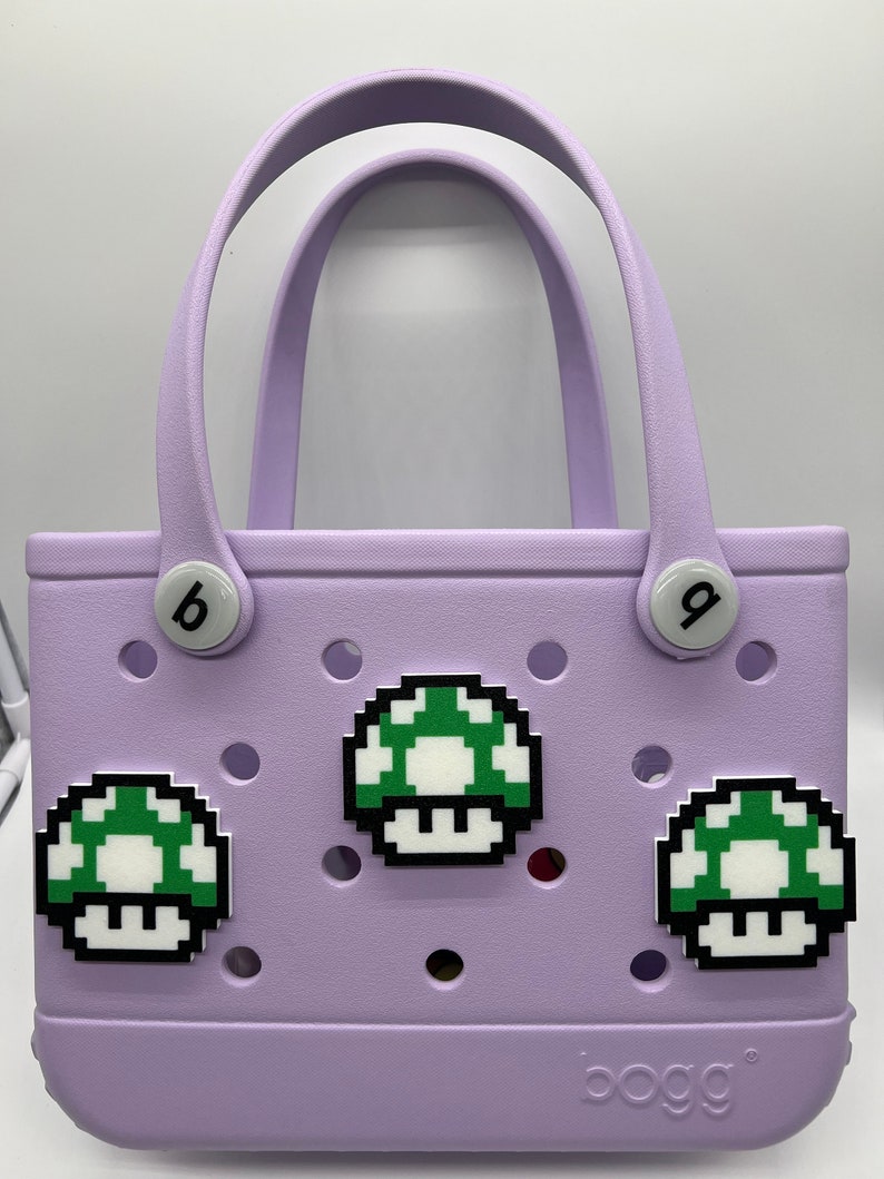Puede incluir: Un bolso tote lavanda con asas blancas y adornos en forma de hongo. La bolsa tiene la palabra "bogg" impresa en la esquina inferior derecha. Tiene dos botones blancos con las letras "b" y "g".