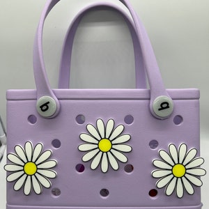 May include: A lavender-colored  Bogg bag with three white daisy flower charms. The bag has a  handle and a  'b' charm on each side.