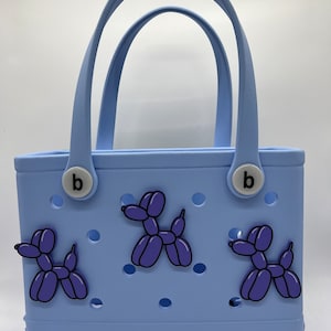 Pode incluir: Uma bolsa tote azul claro com um charme "b" de cada lado. A bolsa tem tr&ecirc;s pingentes de cachorro de bal&atilde;o roxo presos a ela. A bolsa tem a palavra "oog" na parte inferior.