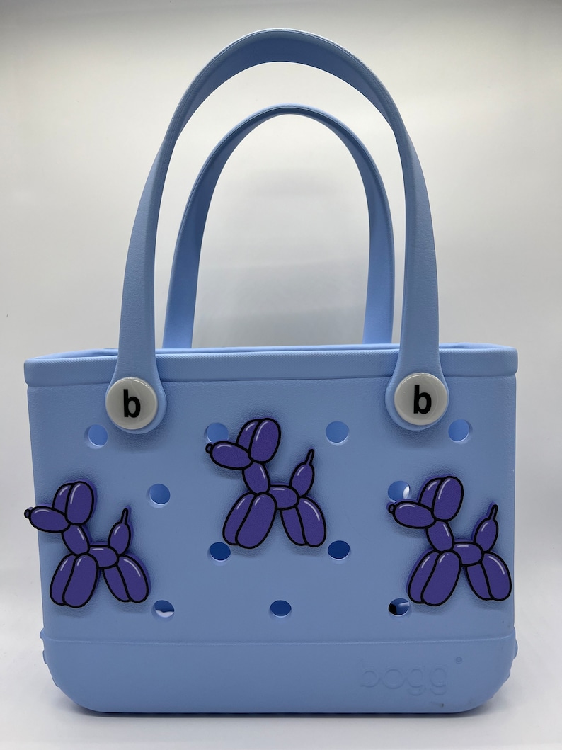 Pode incluir: Uma bolsa tote azul claro com um charme "b" de cada lado. A bolsa tem tr&ecirc;s pingentes de cachorro de bal&atilde;o roxo presos a ela. A bolsa tem a palavra "oog" na parte inferior.
