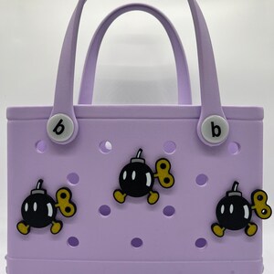 Puede incluir: Un bolso tote lavanda con dos asas y la palabra "bogg" impresa en la parte inferior. La bolsa est&aacute; decorada con tres dijes de bomba de dibujos animados con llaves de cuerda. La bolsa tiene dos botones blancos con una "b" negra.