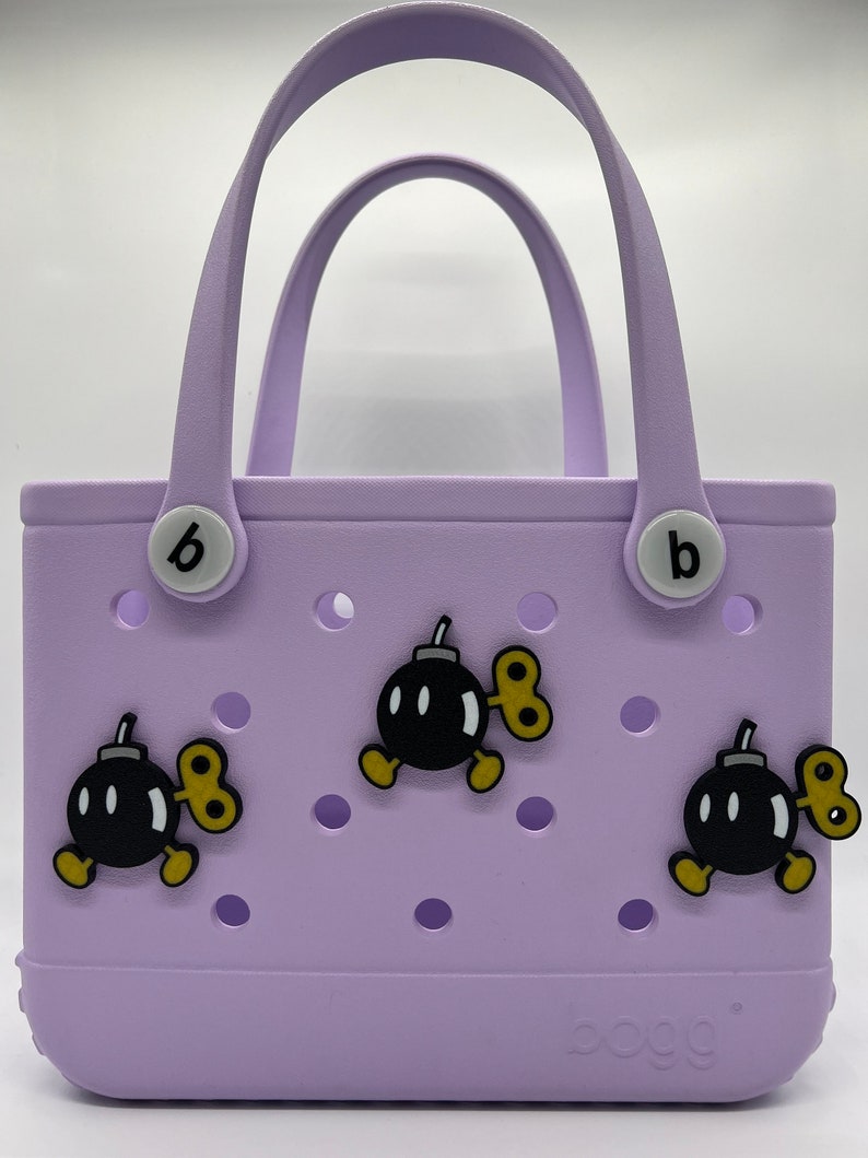 Puede incluir: Un bolso tote lavanda con dos asas y la palabra "bogg" impresa en la parte inferior. La bolsa est&aacute; decorada con tres dijes de bomba de dibujos animados con llaves de cuerda. La bolsa tiene dos botones blancos con una "b" negra.